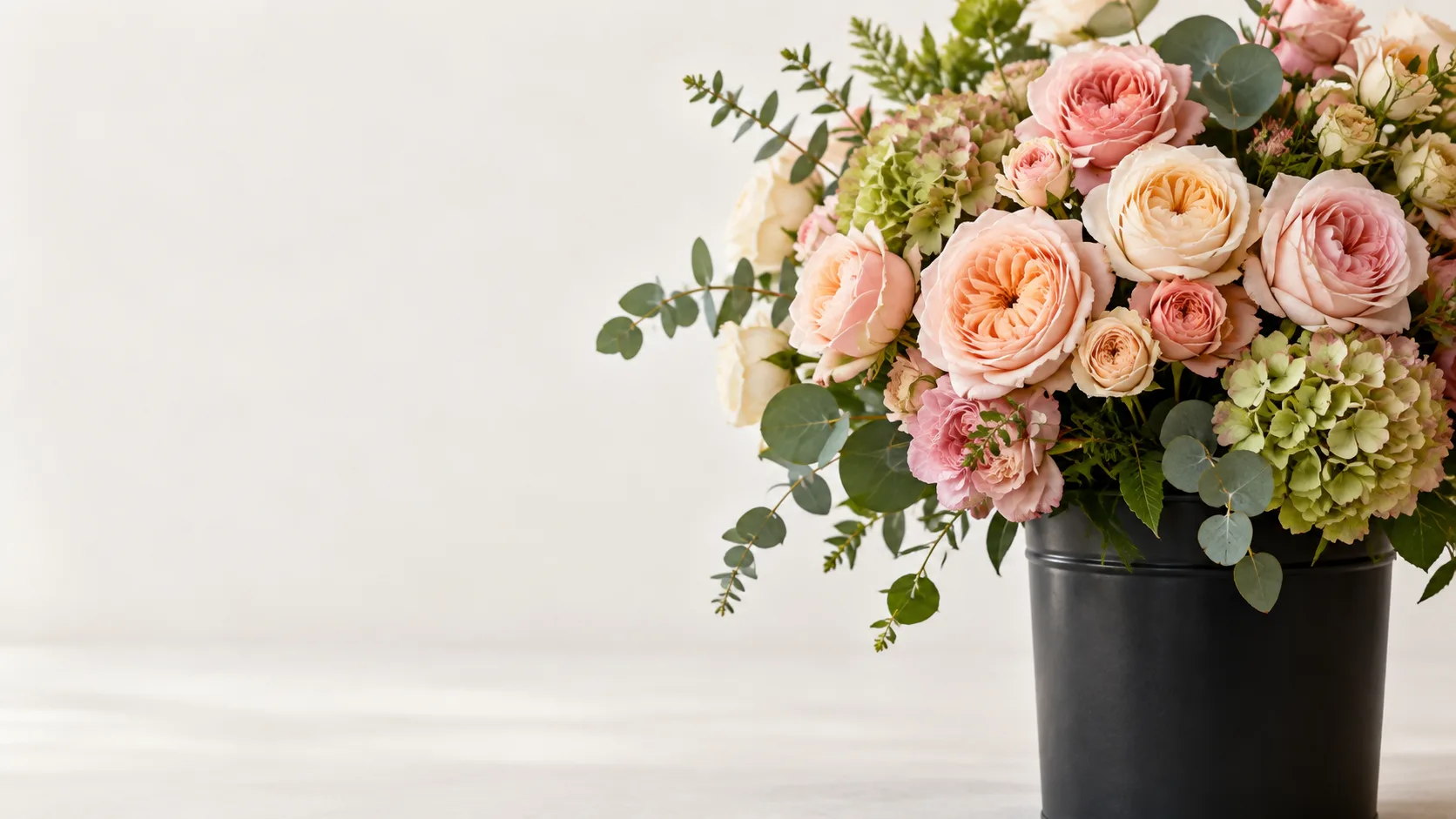 Garden roses in peach and pink tones, green hydrangea and eucalyptus in a black bucket